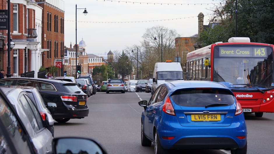 A busy urban street scene featuring parked vehicles lining both sides of the road, including a blue hatchback with a UK registration plate in the foreground, and a red and white Metroline bus displayi